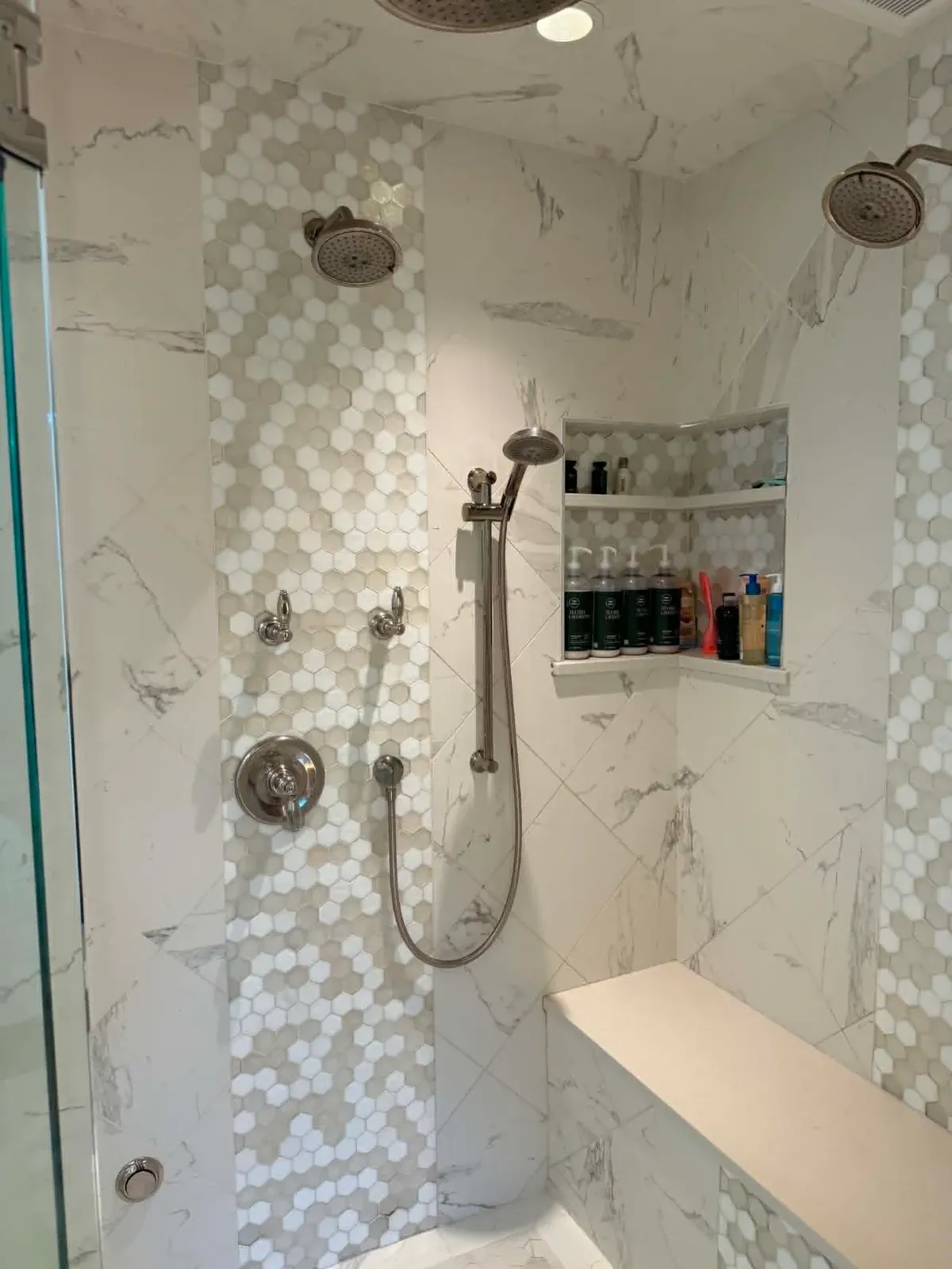 Custom tiled walk-in shower with marble-look walls, hexagon mosaic accent, dual shower heads, and a built-in bench